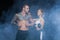 Shirtless muscle man with tattoo and rope posing with beautiful sportswoman holding dumbbell in smoke on black