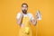 Shocked amazed young man househusband in apron hold in hands iron while doing housework isolated on yellow background