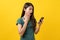 Shocked asian teenager in green tee shirt looking at mobile phone and having shock face on yellow background.