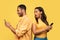 Shocked black woman spying on her boyfriend who is using smartphone, chatting with lover, cheating on yellow background