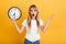Shocked displeased young blonde woman posing isolated over yellow wall background dressed in white casual t-shirt holding clock
