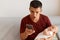 Shocked emotional man with dark hair holding baby girl in hands, using cell phone, looking at device display with scared angry