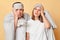 Shocked exhausted couple man and woman in sleep eye mask wrapped in blanket isolated over beige background looking at camera with