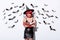 Shocked girl in black witch Halloween costume with red hair holding book near white wall