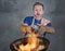Shocked messy man with apron holding pan in fire burning the food in kitchen disaster and unskilled and unexperienced terrible ho