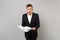 Shocked puzzled young business man in classic black suit white shirt holding paper documents isolated on grey wall