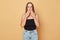 Shocked surprised brown haired young woman wearing black top and jeans standing isolated over beige background looking at