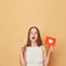 Shocked surprised brown haired young woman wearing casual clothing holding red like icon standing isolated over beige background