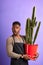 Shocked surprised shopman holding cactus in red pot, looking at camera on purple