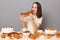 Shocked woman wearing white T-shirt sitting at festive table with various desserts, isolated over gray background holding baked