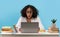 Shocked young black woman sitting at desk, staring at laptop screen, studying or working remotely over blue background