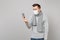 Shocked young man in gray sweater, scarf with sterile face mask looking on mobile phone in hand isolated on grey
