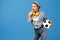 Shoked young woman with yellow penny or skateboard, soccer ball and headphones over blue background.