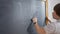 Shooting over shoulder of positive schoolboy drawing on blackboard indoors. Back view close-up of confident Caucasian