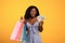 Shopping budget. Portrait of young black woman holding fan of money and colorful shopper bags over orange background