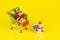 Shopping cart with christmas tree and miniature gift boxes with snowman doll on yellow background. Christmas and New Year shopping