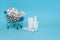 Shopping cart with medicinal pills and thermometer. Spray, vitamins, bottle lying near mini basket on blue background. Pharmacy