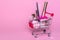 Shopping trolley with makeup on a pink background. Perfume, brush, mascara