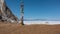 On the shore of a frozen lake, on bare ground, there is a wooden ritual pillar tied with bright multi-colored ribbons.