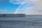 A shot of a brown wooden pier surrounded by vast blue ocean water with a blue sky, clouds and a rainbow in the sky