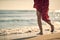 Shot of legs of young woman in red summer dress walking on beach by waves of water. Fashion, summertime, lifestyle concept