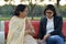 Shot of a senior retired Indian woman sitting in a park with her daughter in law on a red bench peeling peanuts, and eating them.