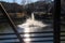 A shot of a water fountain in the center of a rippling green lake in the park surrounded by yellow winter grass, bare winter