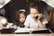 Siblings lie in hut of chairs and blankets. Brother and sister reading book with a flashlight at home