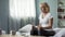 Sick blond lady sitting on bed with glass of water and pills in front, treatment