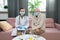 Sick elderly patient and young general practitioner in masks sitting on couch
