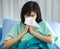 A sick female patient lean on a pillow, sitting on bed catching a cold or flu, coughing and sneezing into facial tissue severely.