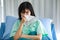 A sick female patient lean on a pillow, sitting on bed catching a cold or flu, coughing and sneezing into facial tissue severely.