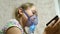 A sick girl in a mask inhales a spray bottle and plays a computer game on a tablet. Kid in a blue hospital mask inhale