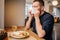 Sick man is sitting at the table and sneezing. He is covering his nose with white napkin and keeping eyes closed. Man is