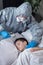 A sick school-age boy is lying in bed. A person wearing a protective suit and rubber gloves measures the child`s temperature with