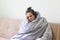 Sick young woman in blanket sits at home with severe headache