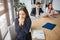 Sick young woman sit saparately at table in meeting room and sneezing. She hold white napkin. Her colleagues work behind.