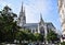 Side of a large Gothic church whose spiers rise above the trees in the blue sky stained by white clouds.