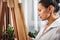 Side portrait of a beautiful woman painter wearing a white shirt and standing in a front of easel