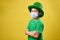 Side portrait of a boy in green Irish hat and protective medical mask poses to camera with arms crossed. Saint Patrick day