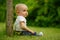 Side portrait of little cute baby in jeans sitting on grass near tree trunk in garden, closeup, bright green background