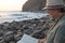 Side view of alone senior woman with  hat sitting on the beach of pebbles reading a book. Waves of the ocean. Sunset light