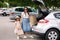 Side view of attractive young mom with her adorable little daughter stand by open trunk with shopping bag