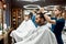 Side view of a barber girl making new trendy haircut for a young handsome bearded man sitting in barbershop chair