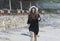 Side view of barefooted young ethnic woman in black dress and hat walking on sandy beach during summer vacation in tropical resort