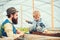 Side view bearded man looking at lovely kid playing in box full of soil. Daddy in blue vest, yellow shirt and hat