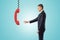 Side view of businessman standing and reaching out for big red landline phone receiver dangling down on wire from above.