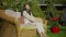 Side view of charming slim Asian woman stretching sitting on cozy couch in sunny garden looking around smiling. Wide