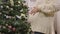 Side view of female Caucasian hands caressing pregnant belly with decorated Christmas tree at background indoors