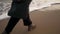 Side view of female feet in boots walking along sea waves foam on sandy beach. Woman in green coat goes along ocean